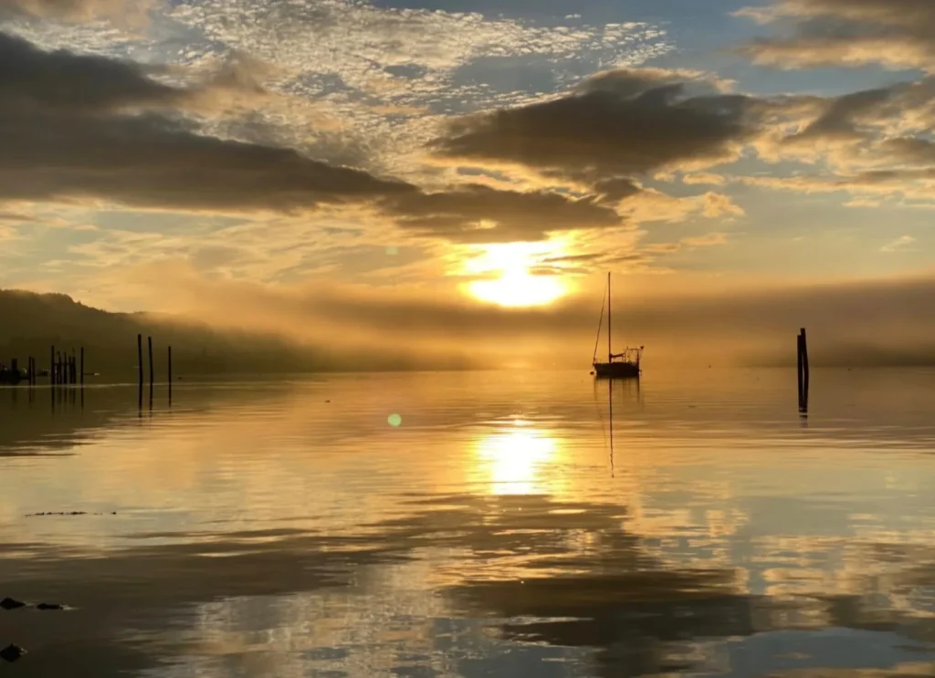 Ein Boot auf einem stillen See im Morgenlicht, dahinter die Silhouette der Berge