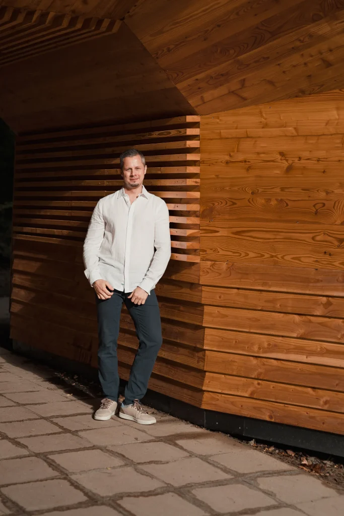 Max Simon steht in weißem Hemd vor einer Wand aus waagerechten Holzlatten im Abendlicht
