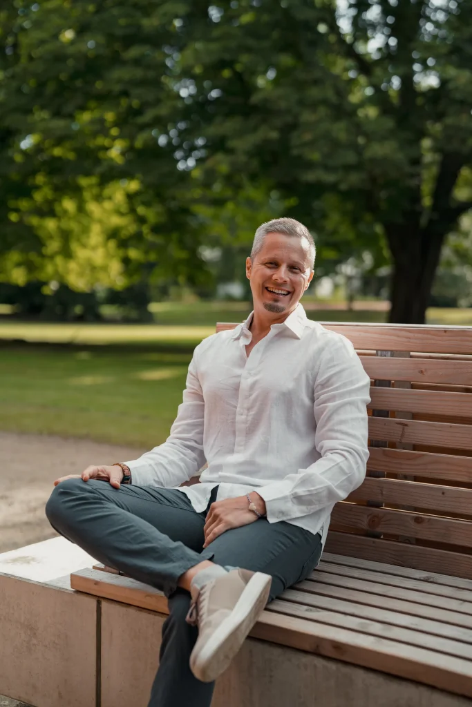 Max Simon sitzt mit übereinandergeschlagenen Beinen auf einer hölzernen Parkbank, im weißen Leinenhemd, und lacht in die Kamera